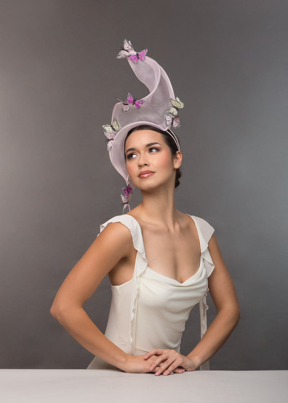 A model wearing a decorative pale pink crinoline headpiece with butterflies against a grey background